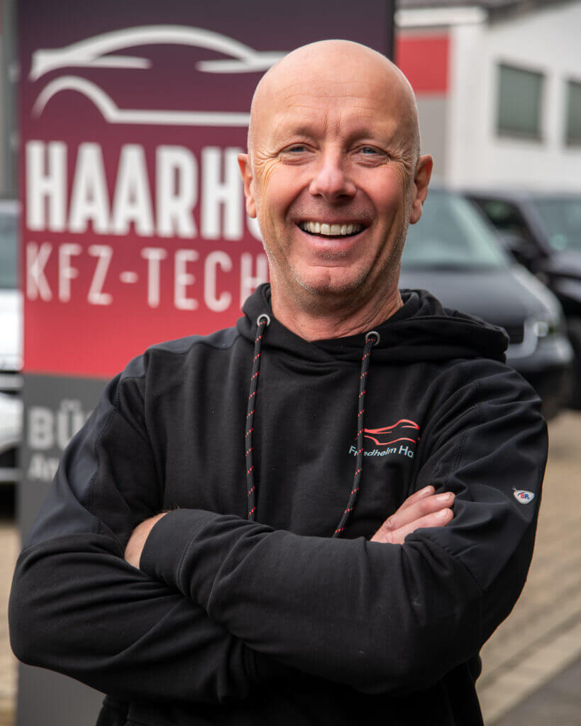 Geschäftsführer vor dem Firmenlogo der KFZ-Technik Haarhoff, lächelnd in schwarzem Kapuzenpullover mit verschränkten Armen. Im Hintergrund ist das bordeauxrote Firmenschild und Teile des Werkstattgebäudes zu sehen.